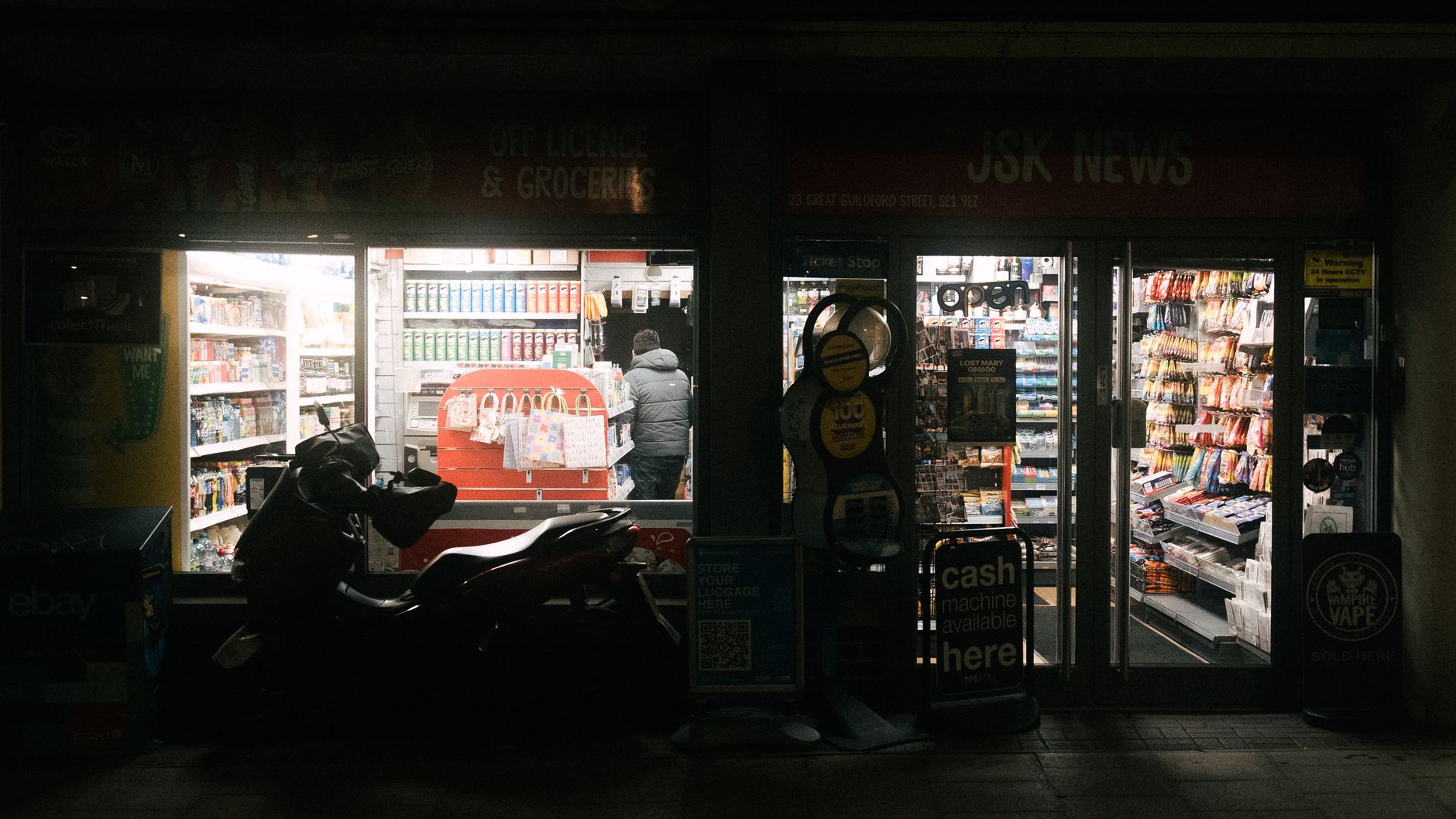 Petite supérette éclairée la nuit, rayons de boissons et de snacks visibles derrière la vitrine, deux-roues garé devant