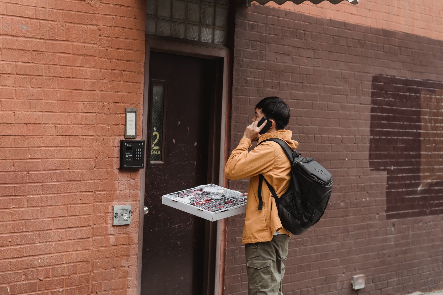 Livreur avec un sac isotherme sur le dos, boîtes à pizza en main, qui téléphone devant la porte d'un immeuble
