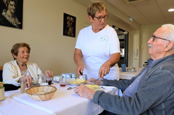 Résidents âgés attablés au restaurant servis par une aide-soignante souriante